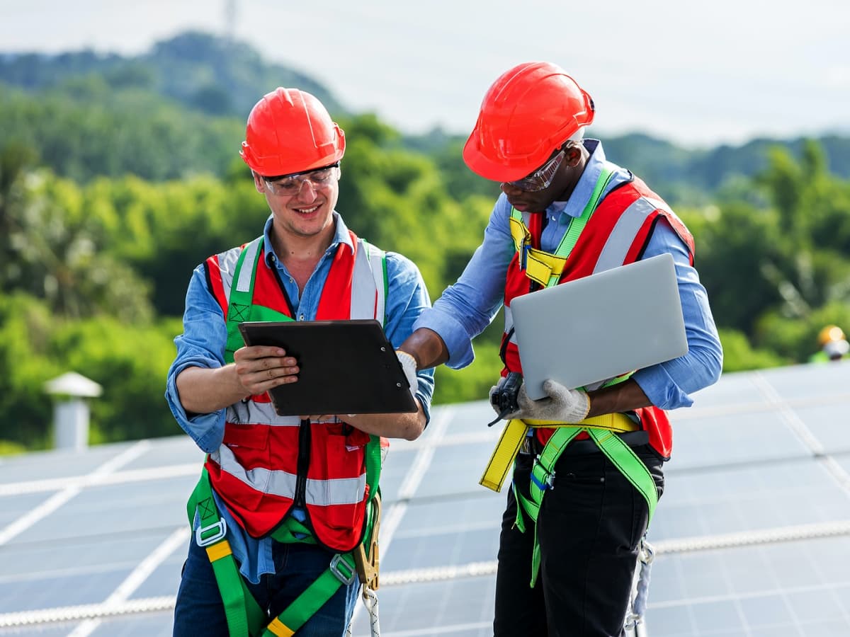 Two men in safety vests and hard hats on a rooftop, working on a laptop.